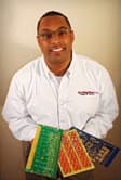 A smiling African-American male with neatly trimmed hair and 👓 wearing business casual clothing including a white button shirt with 'the component store' logo imprinted on it and holding a collection of circuit boards
